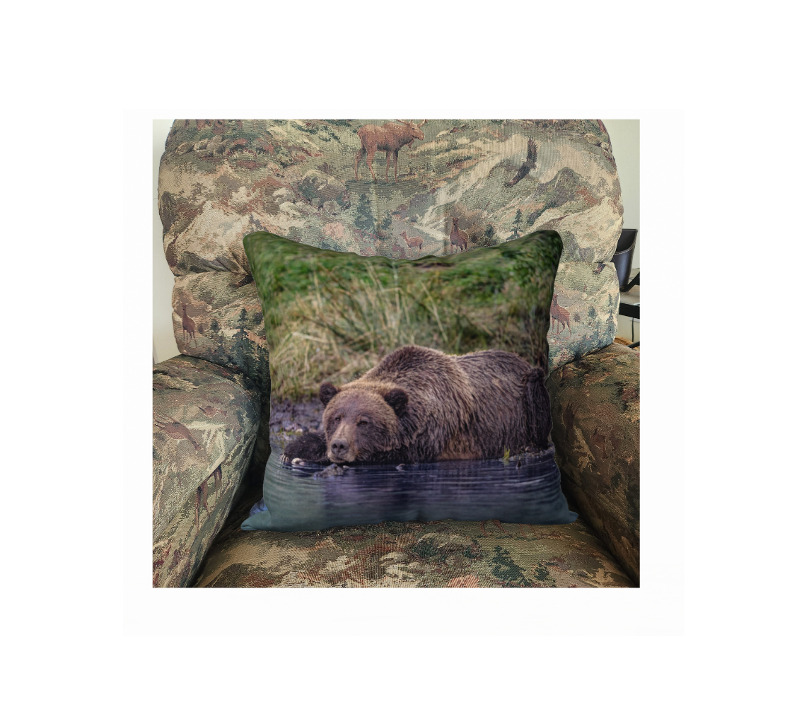 This 18 x 18 inch plush velveteen cushion cover features a photo of a big brown female grizzly bear at rest in the water along the river's edge. The cushion cover background is various tones of tall green grass, and the water in which the bear is lying in is a dark blue. This image is of a cushion cover on a pillow form in a chair. 