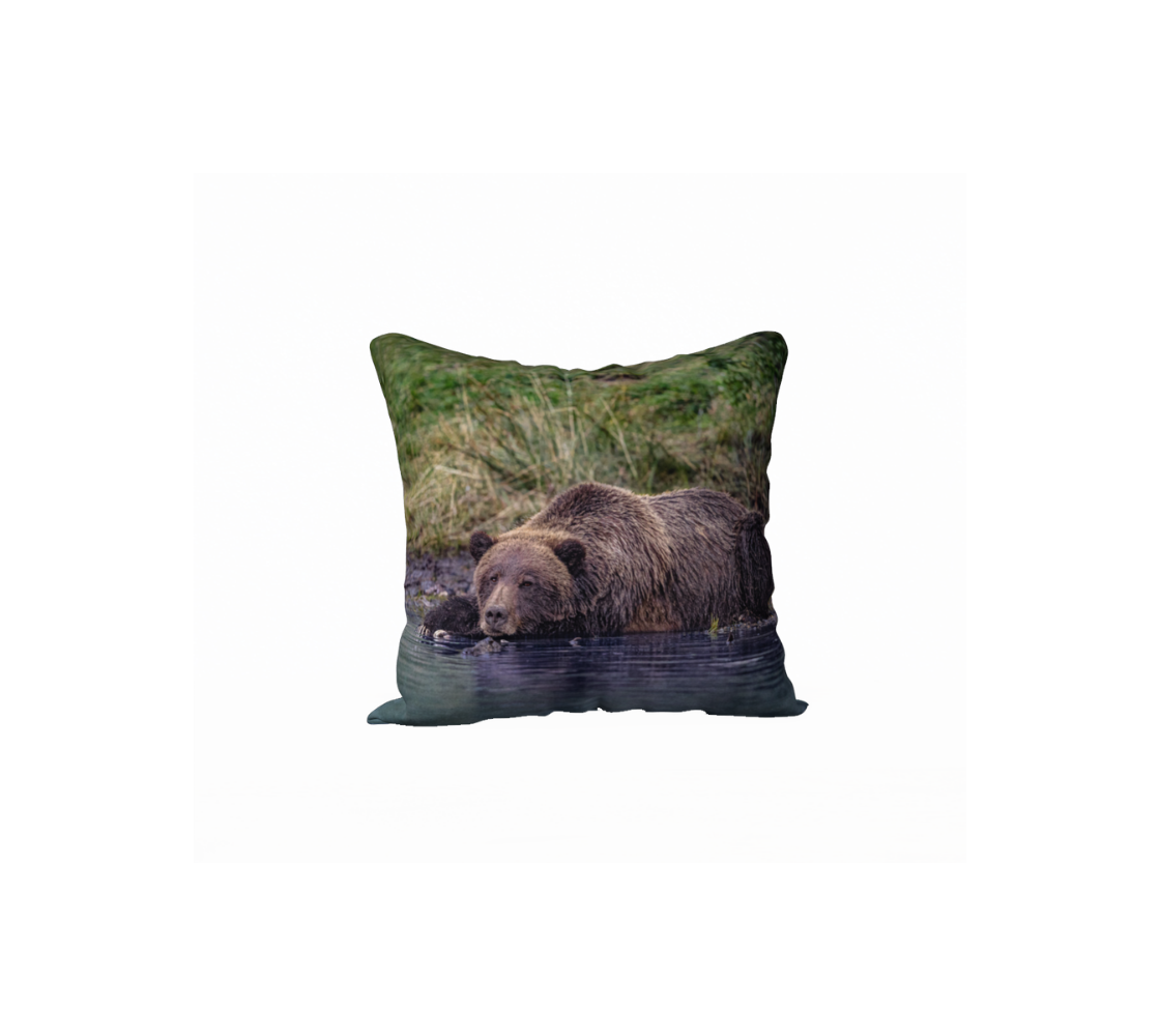 This 18 x 18 inch plush velveteen cushion cover features a photo of a big brown female grizzly bear at rest in the water along the river's edge. The cushion cover background is various tones of tall green grass, and the water in which the bear is lying in is a dark blue.