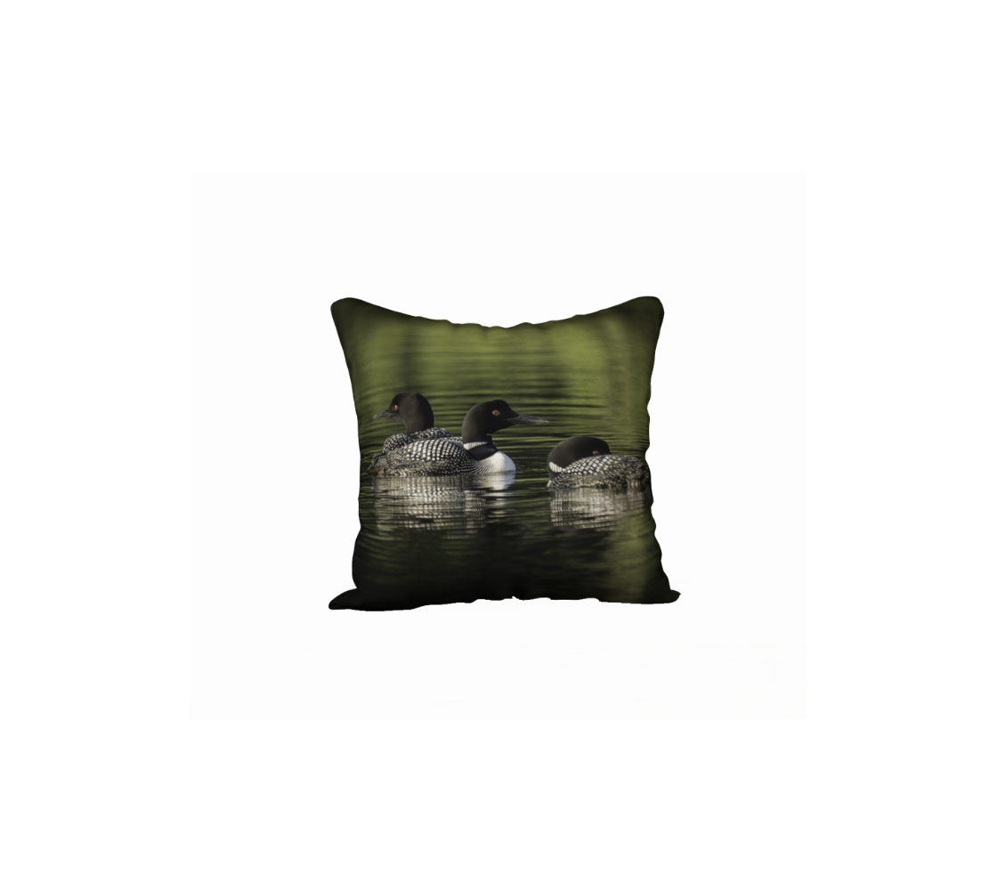 This 18 x 18 inch plush velveteen cushion cover features a photo of three black and white loons with red eyes, at rest on a green colored lake. The cushion cover background is various blurred green tones due to the reflection of the evergreens around the lake.