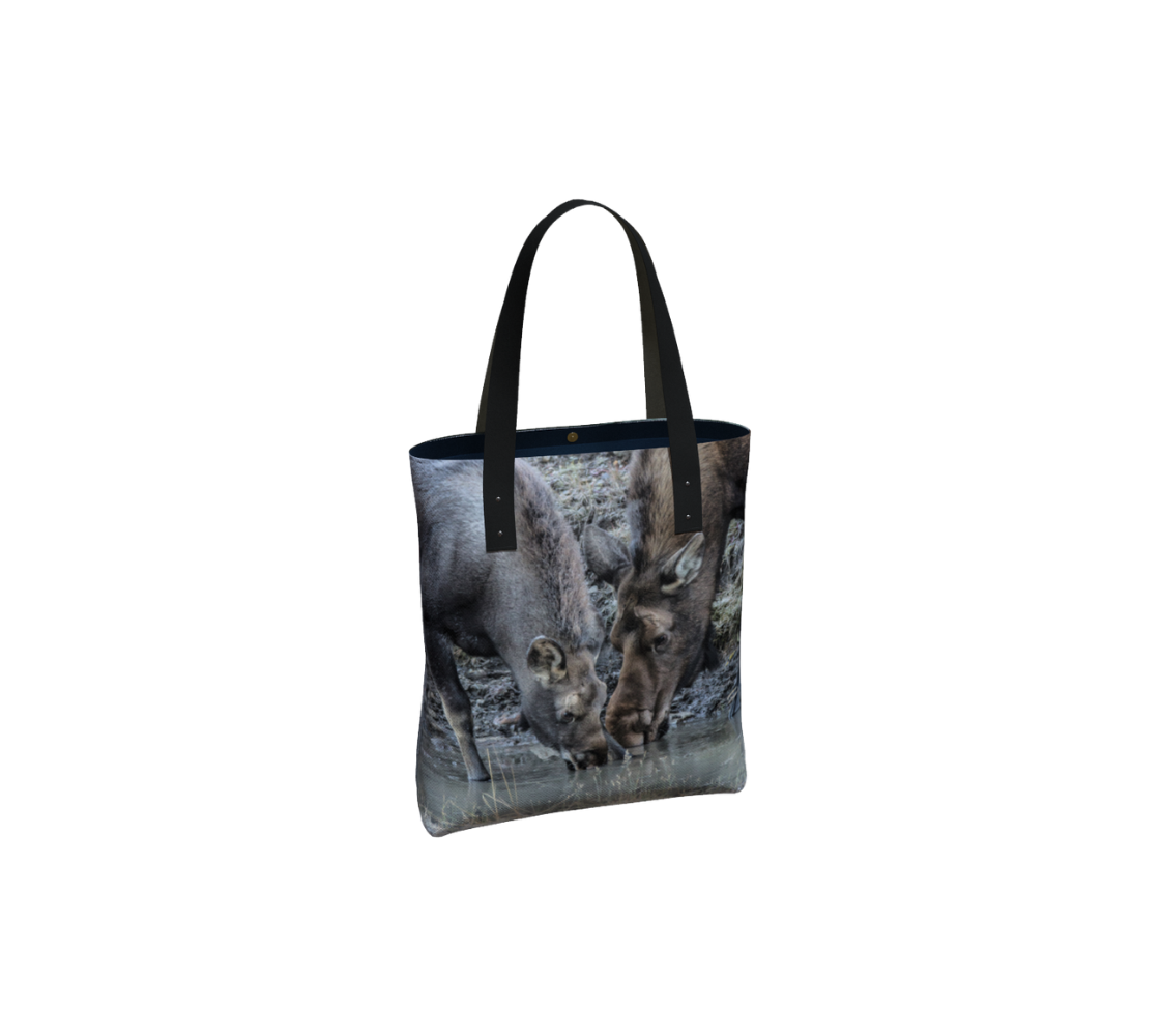 This canvas tote bag has an image of a cow and calf moose head and shoulders, stretched down drinking water from the ditch. The background is grey and blue rocks and grass, due to the morning frost on the ground. It has a one inch wide black vegan shoulder strap with navy blue lining.