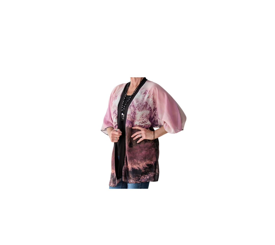 Tthis frontview is of a lady wearing a semi-sheer poly chiffon kimono robe featuring a photo of a mountain scene . The background is a pink mountain sunset scene, with pine trees and a creek running along the bottom. Kimono has black lightweight bamboo trim.