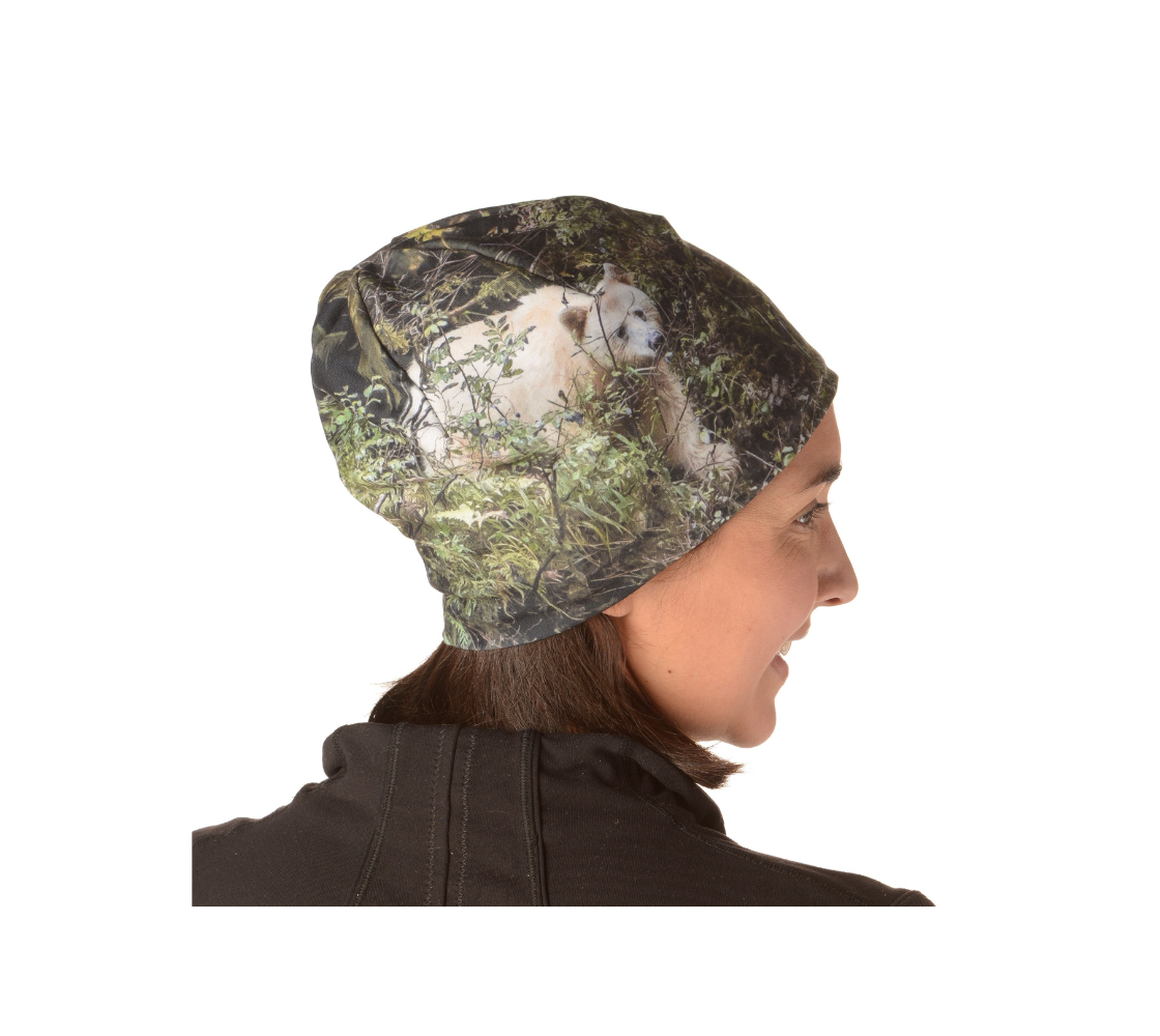 This is a side portrait of a lady wearing a photo of a spirit bear on a beanie. The background is green bushes and brown branches of the Great Bear Rainforest. The white spirit bear is stepping down out of the forest and is making her way to the stream below.