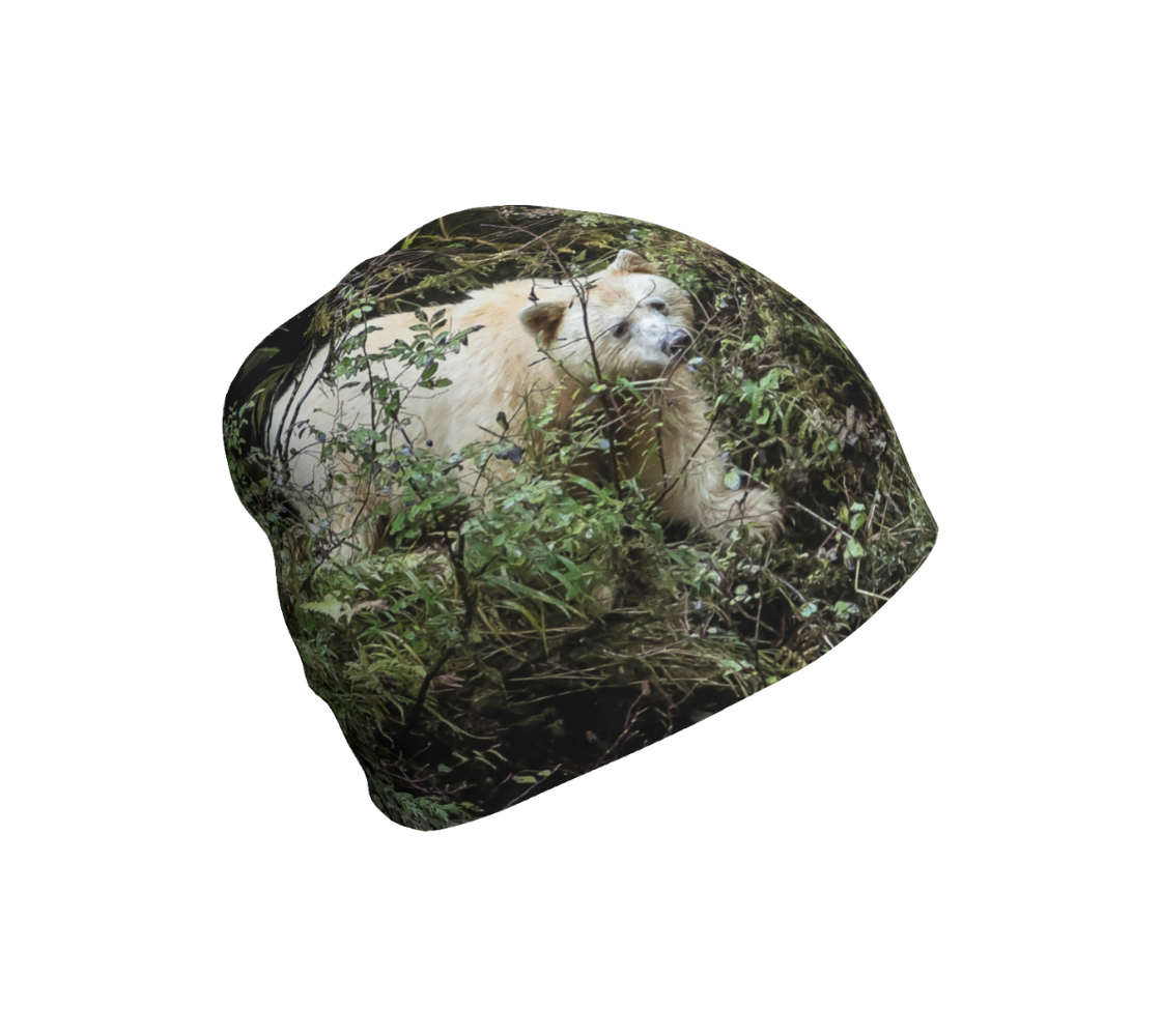 This is a photograph of a spirit bear on the right side of a beanie. The background is green bushes and brown branches of the Great Bear Rainforest. The white spirit bear is stepping down out of the forest and is making her way to the stream below.