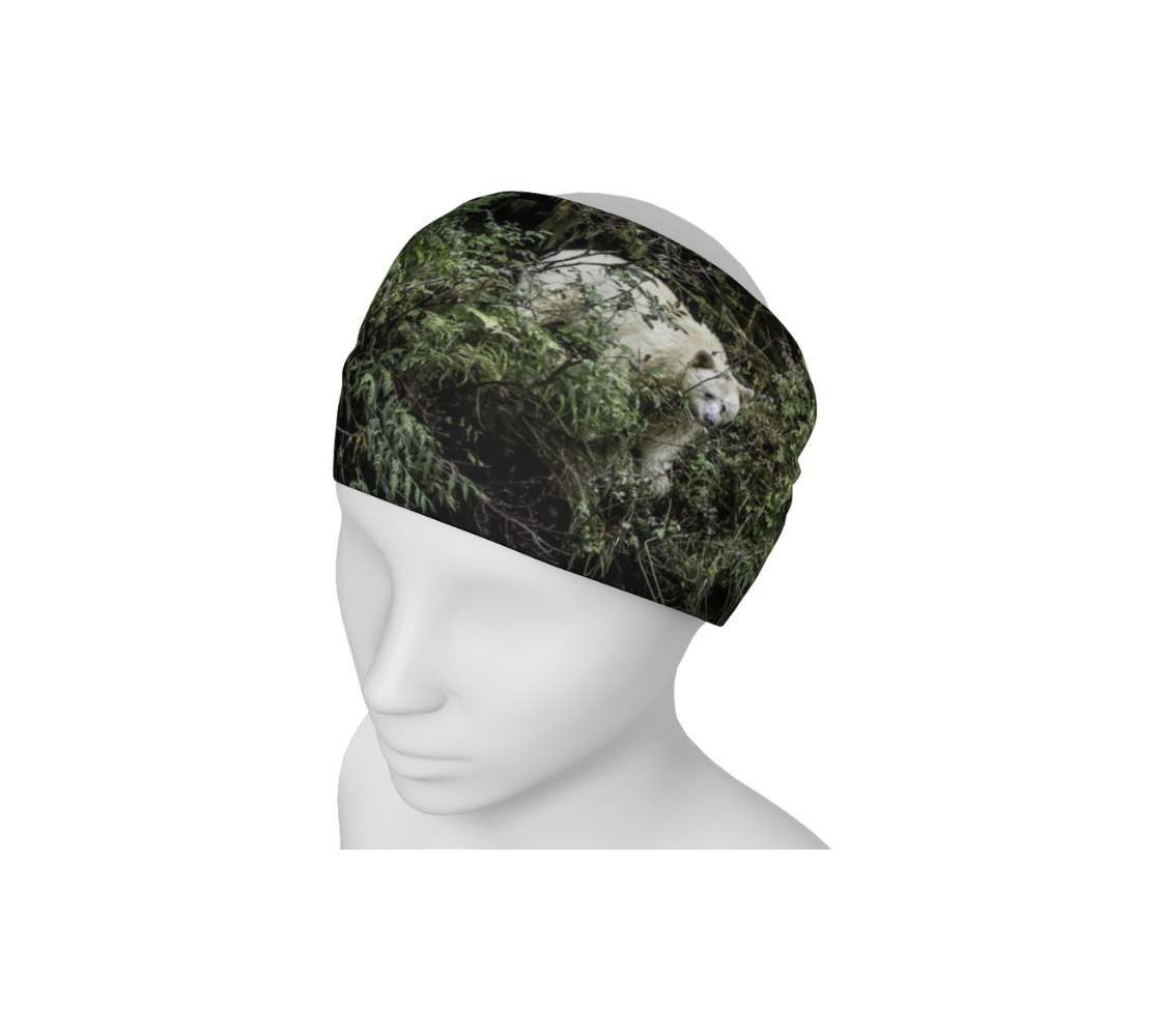 This is an image of a photo of a spirit bear on a head and neck band on a mannequin worn as a headband. The background is green bushes and brown branches of the Great Bear Rainforest. The white spirit bear is stepping down out of the forest and is making her way to the stream below.