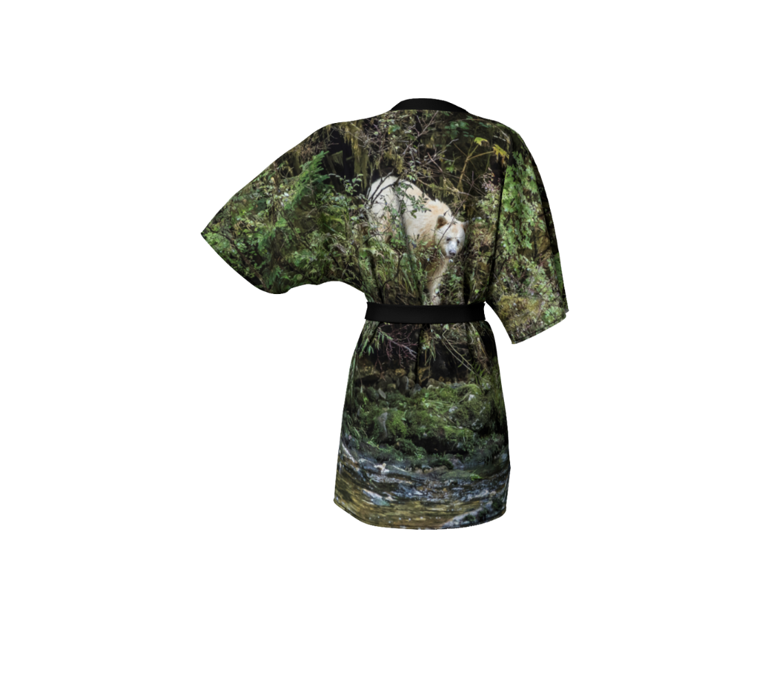 This backview is a semi-sheer poly chiffon kimono robe featuring a photo of a spirit bear. The background is of green trees and moss with a white spirit bear coming down a creek bank into the water. Kimono has black trim and a black lightweight bamboo belt for tying.