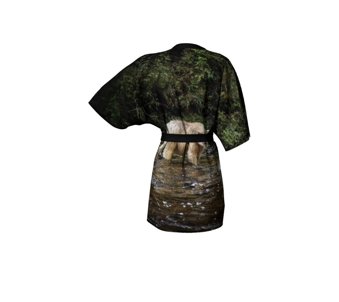 This backview is a semi-sheer poly chiffon kimono robe featuring a photo of a spirit bear. The background is of green trees and moss with a gold and green colored creek.  The featured image in the middle is a white spirit bear walking in the stream looking for fish. Kimono has black trim and a black lightweight bamboo belt for tying.