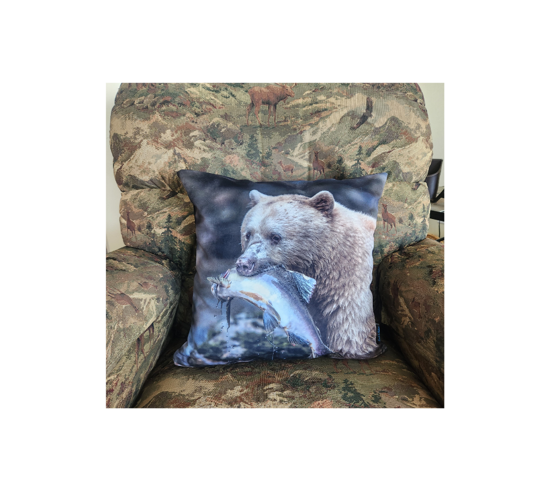 This 18 x 18 inch plush velveteen cushion cover features a photo of a white spirit bear holding a light grey salmon in her mouth. The background is mottled green and grey tones with a mossy rock. This image is of the cushion cover on a pillow form in a chair.