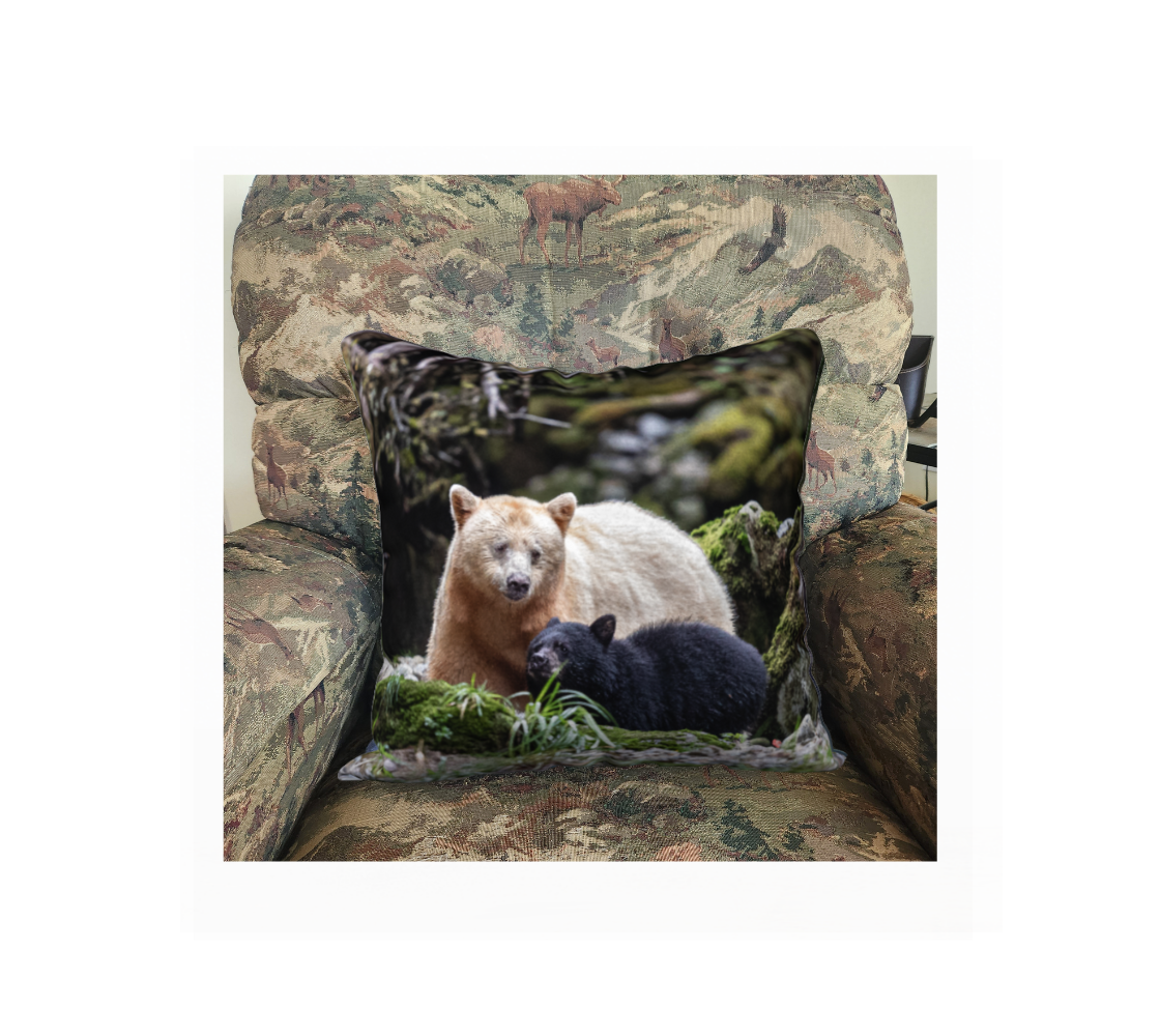 This 18 x 18 inch plush velveteen cushion cover features a photo of a white momma spirit bear with her little black cub beside her. The cushion cover background is green moss and grass wih grey rocks and branches. This image is of a cushion cover on a pillow form in a chair.