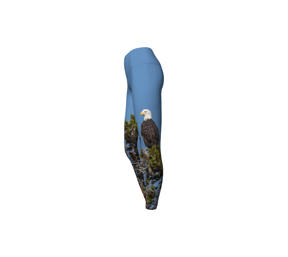 This is a side profile of a pair of yoga leggings featuring a bald eagle on the side of each leg, with a 4.5 inch high waist band. The background is a blue sky with a bald eagle perched on top of a pine tree. 