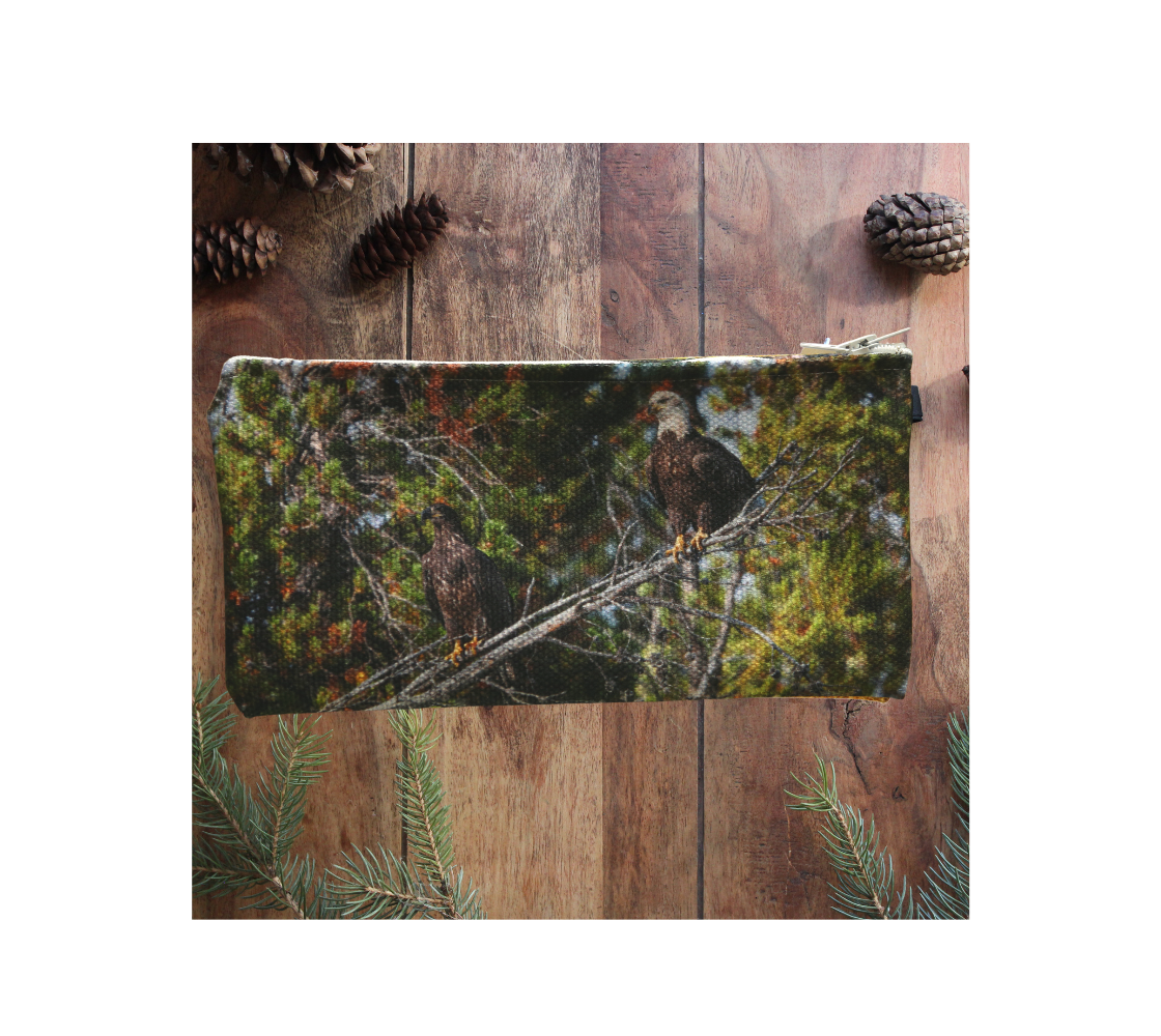 This durable double sided 9” x 4” canvas zippered pouch features 2 real images of bald eagles. The background is of trees and branches in various tones of green and grey. There are 2 bald eagles, one is a juvenile and the other is an adult, that are perched on a  grey tree branch