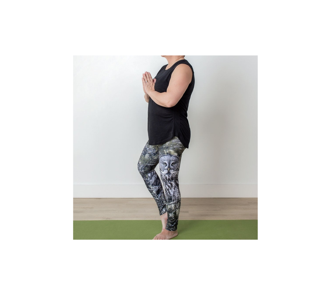 This is a side profile of a lady wearing a pair of yoga leggings with an owl on the side of each leg. The background is green pine boughs and lichen hanging from the grey pine tree branches, with a great grey owl with black, grey and white feathers, and yellow eyes, perched on a branch. 