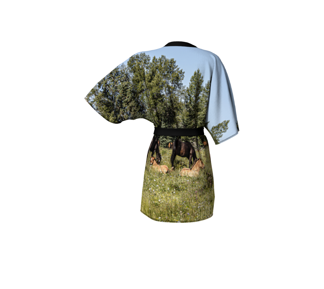 This backview is a semi-sheer poly chiffon kimono robe featuring a photo of wild horses. The background is  of a blue sky, green trees and grass with wild foals lying among the wildflowers and many wild horses grazing in the field. Kimono has black trim and a black lightweight bamboo belt for tying.