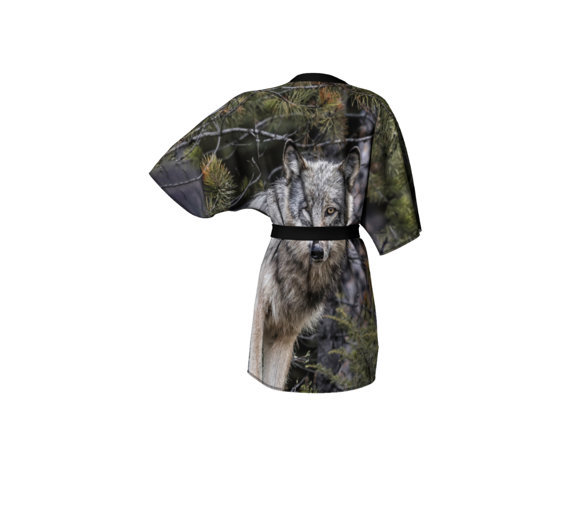 This backview is of a semi-sheer poly chiffon kimono robe featuring a photo of a wolf. The background is green and grey with pine tree branches with a head shot of a grey wolf. Kimono has black trim and a black lightweight bamboo belt for tying.